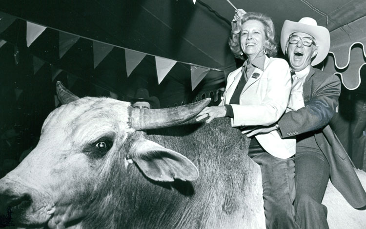 Jane y John Justin se ríen mientras posan sobre un toro bravo para una foto. 
